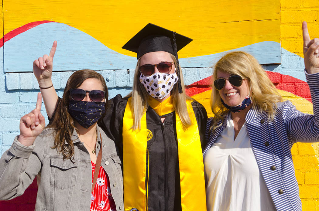 An M-S-W graduate and two guests make the No. 1 sign with their index fingers in front of a wall painted yellow and light blue. All are wearing masks.