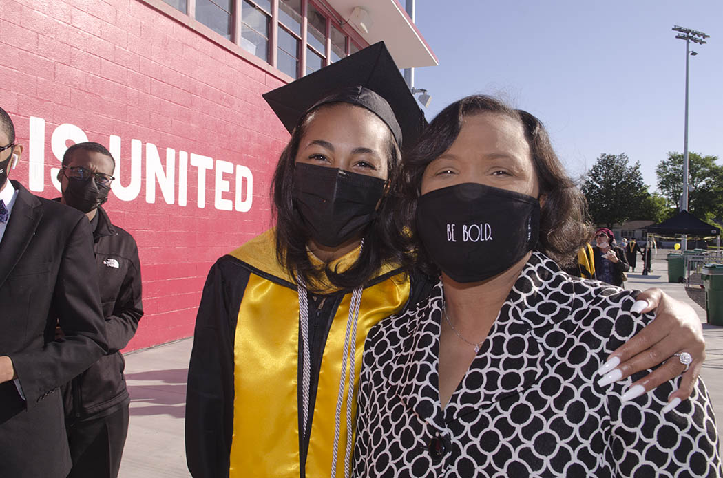 An M-S-W graduate puts their arm around a guest, whose mask reads: Be Bold. Additional guests stand to the side in front of a red-painted building