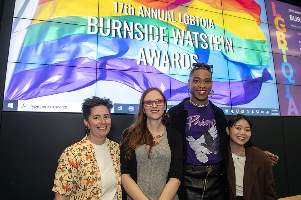 Four people standing in front of a screen with a rainbow flag and white text that reads 17th ANNUAL LGBTQIA BURNSIDE WATSTEIN AWARDS