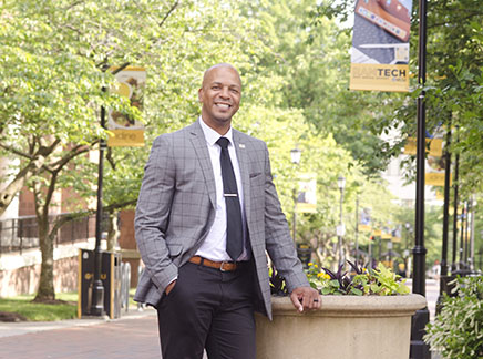 Social work alum Anthony Estreet, wearing a gray sports coat, dark tie and pants, stands on the VCU campus, resting against a large flower planter.