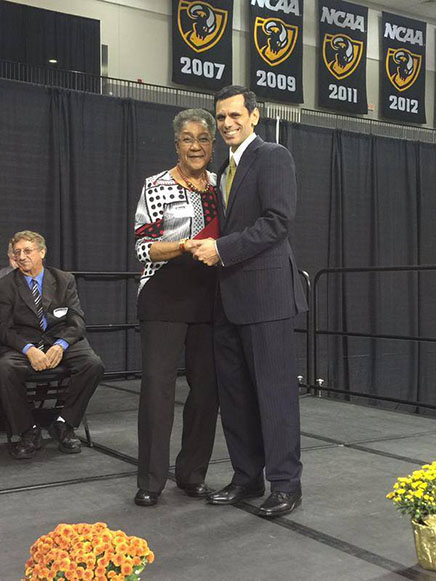 Lila Garlick standing on a stage and receiving an award from VCU President Michael Rao. Black and gold NCAA basketball banners from 2007, 2009, 2011 and 2012 hang in the background.