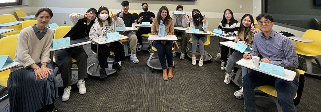 A group of visiting South Korean students sit in yellow desk chairs around social work associate Traci Wike in a classroom. Each student has a blue card with their name sitting on their desks.