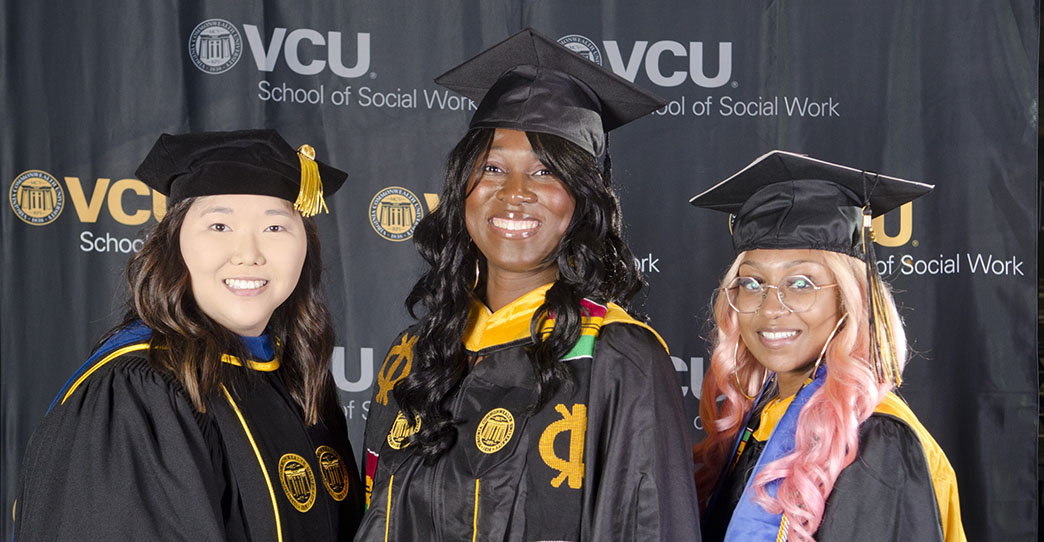 Three students in graduation regalia stand in front of a black backdrop printed with VCU School of Social Work.