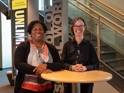 Two people stand with their hands resting on a table. A sign in the background reads Welcome to the School of Social Work.