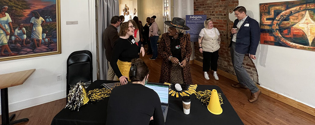 A group of people stand in front of a registration table that has black and gold pompoms and a gold megaphone. On the wall are two colorful pieces of art, and part of the wall is expose brick.