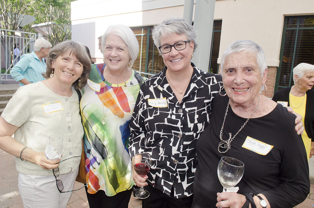 Four women stand together with their arms around each other, one in a light top, one in a multi-colored top, one in a black and white-patterned top and one in a black top.