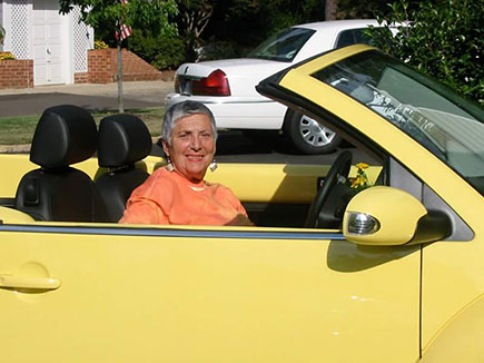 Flossie Segal sits in a yellow Volkswagen Beetle convertible, smiling.