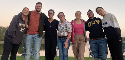 A group of seven people stand arm in arm in front of a lake near sunset.