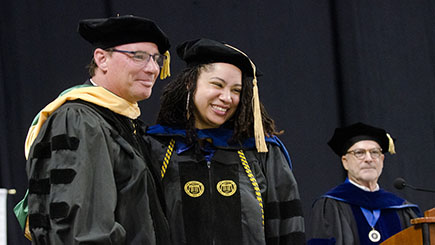 Two people stand together and smile while a third looks on in the background. All three are wearing academic regalia of black caps and gowns.