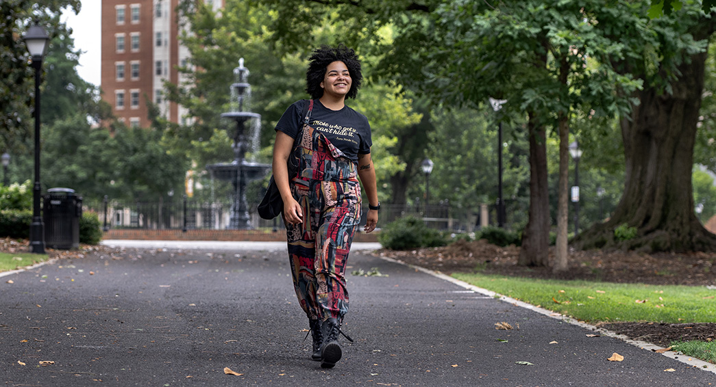 Jae Lange walks down a paved pathway in a park with the backdrop of trees and a fountain.