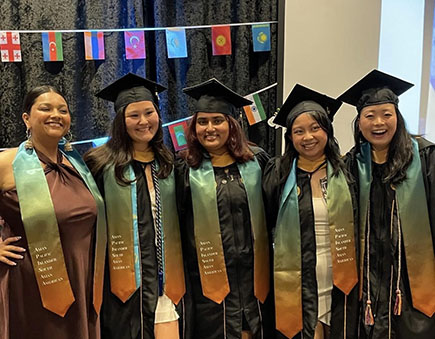 A group of five graduates stands arm in arm with stoles representing them as Asian Pacific Islander or South Asian American graduates.