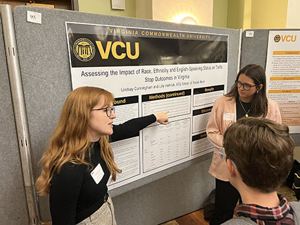 Two student presenters gesture in talking about their research poster to an attendee during a research symposium.