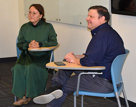 Two people sit at desks, one in a green dress and one in a blue shirt and dark pants.