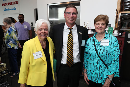 Three people stand together smiling, one in a yellow jacket, one in a suit and one in a blue-green blouse with black stripes.